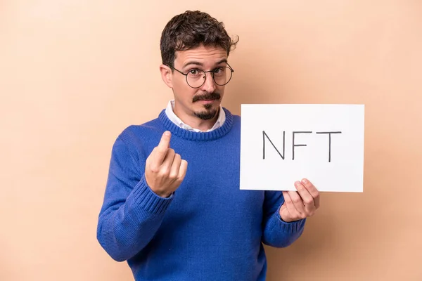 Young caucasian man holding a NFT placard isolated on beige background pointing with finger at you as if inviting come closer.