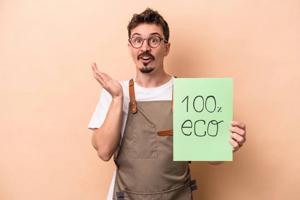Young caucasian gardener man holding 100% eco placard isolated on beige background surprised and shocked.