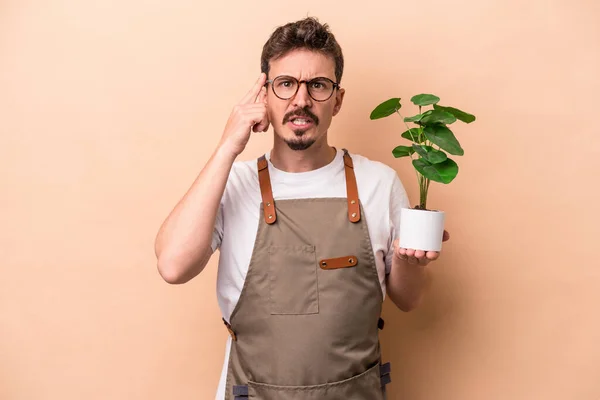 Young caucasian gardener man holding a plant isolated on beige background showing a disappointment gesture with forefinger.