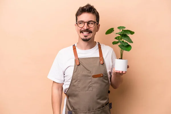 Młody Biały Ogrodnik Człowiek Trzyma Roślinę Odizolowany Beżowym Tle Patrzy — Zdjęcie stockowe