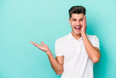 Mavi arka planda izole edilmiş genç beyaz bir adam avucunun içinde fotokopi alanı tutuyor, el üstünde tutuyor. Şaşırmış ve mutlu..