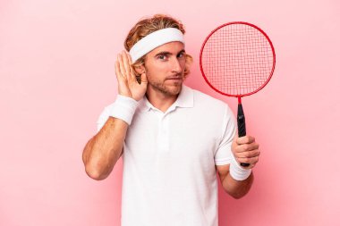 Badminton oynayan beyaz bir adam pembe arka planda izole edilmiş bir şekilde bir dedikoduyu dinlemeye çalışıyor..