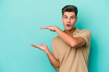 Mavi arka planda izole genç beyaz adam şok ve hayret içinde elleri arasında bir kopya boşluk tutuyor.