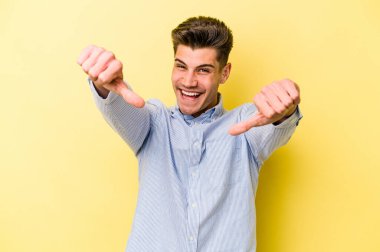 Sarı arka planda izole edilmiş genç beyaz adam iki parmağını da kaldırıyor, gülümsüyor ve kendinden emin..