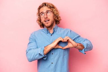 Pembe arka planda izole edilmiş genç beyaz adam gülümsüyor ve elleriyle kalp şeklini gösteriyor..