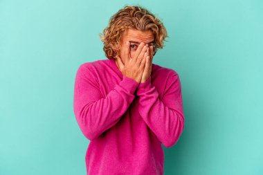 Mavi arka planda izole edilmiş genç beyaz adam kameraya parmaklarıyla göz kırpıyor, yüzünü kapatmaktan utanıyor..