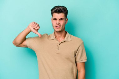 Mavi arka planda izole genç beyaz adam baş parmağını aşağı gösteriyor, hayal kırıklığı konsepti.