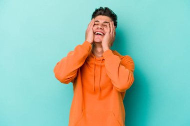 Mavi arka planda izole edilmiş genç beyaz adam ellerini başının üstünde tutarak neşe içinde gülüyor. Mutluluk kavramı.