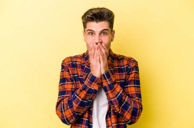 Sarı arka planda izole edilmiş genç beyaz adam elleriyle ağzını kapatırken şok oldu..