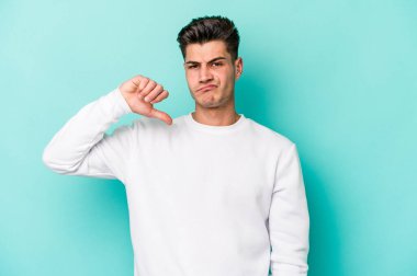 Mavi arka planda izole genç beyaz adam baş parmağını aşağı gösteriyor, hayal kırıklığı konsepti.