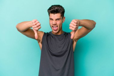 Mavi arka planda izole edilmiş genç beyaz bir adam baş parmağını aşağı gösteriyor ve hoşlanmadığını ifade ediyor..