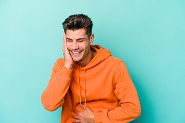 Mavi arka planda izole edilmiş genç beyaz adam mutlu bir şekilde gülüyor ve ellerini karnının üstünde tutarak eğleniyor..