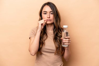 Beyazlı genç bir kadın bej arka planda izole edilmiş bir şişe su tutuyor ve parmakları dudaklarında bir sır saklıyor..