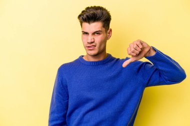 Sarı arka planda izole edilmiş genç beyaz adam baş parmağını aşağı gösteriyor, hayal kırıklığı konsepti..