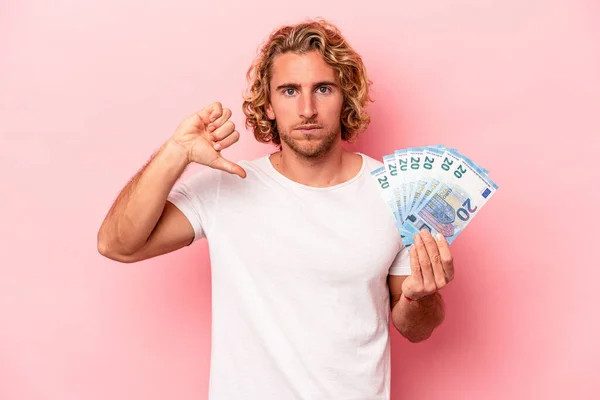 Young caucasian man holding banknotes isolated on pink background showing a dislike gesture, thumbs down. Disagreement concept.