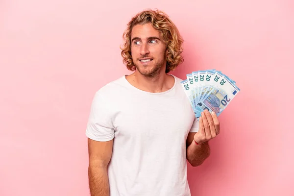 Young caucasian man holding banknotes isolated on pink background looks aside smiling, cheerful and pleasant.