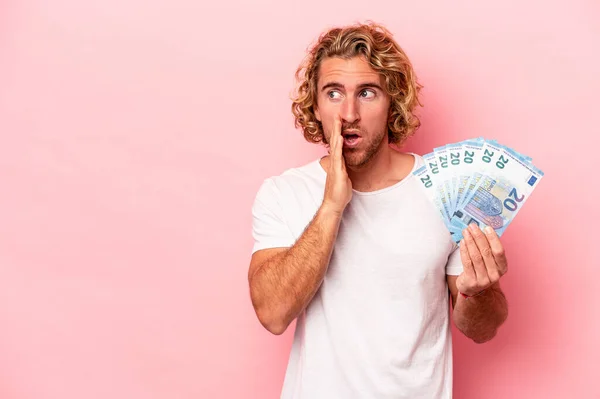 Young caucasian man holding banknotes isolated on pink background is saying a secret hot braking news and looking aside