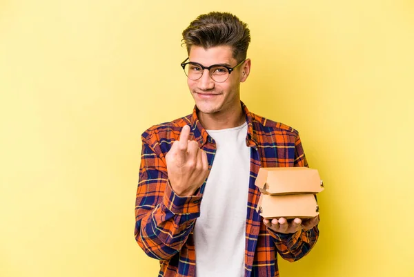 Joven Caucásico Sosteniendo Una Hamburguesa Aislada Sobre Fondo Amarillo Señalándote — Foto de Stock