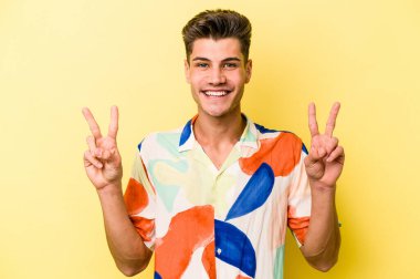 Sarı arka planda izole edilmiş genç beyaz adam zafer işareti gösteriyor ve gülümsüyor..