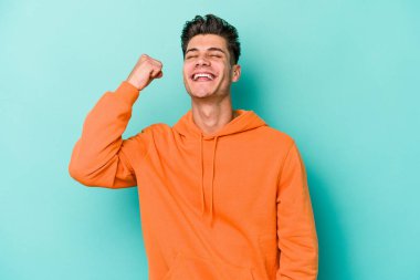 Mavi arka planda izole edilmiş genç beyaz bir adam zaferi, tutkuyu ve coşkuyu kutluyor, mutlu bir ifade..