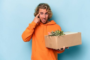 Young caucasian man making a move while picking up a box full of things isolated on blue background showing a disappointment gesture with forefinger.
