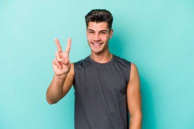 Mavi arka planda izole edilmiş genç beyaz adam zafer işareti gösteriyor ve gülümsüyor..