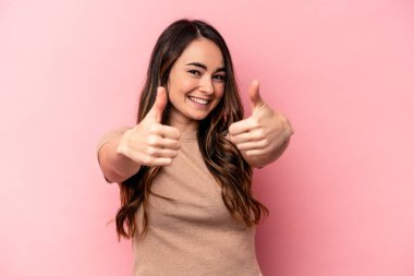 Pembe arka planda izole edilmiş genç beyaz kadın iki parmağını da kaldırıyor, gülümsüyor ve kendinden emin..