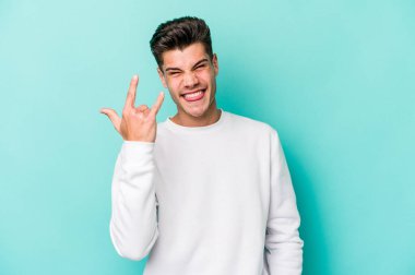 Mavi arka planda izole edilmiş genç beyaz adam parmaklarıyla kaya hareketi gösteriyor.