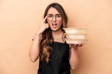 Beyaz tenli genç bir kadın bej arka planda saklanmış vaziyette işaret parmağıyla yaptığı hayal kırıklığı jestini gösteriyor..