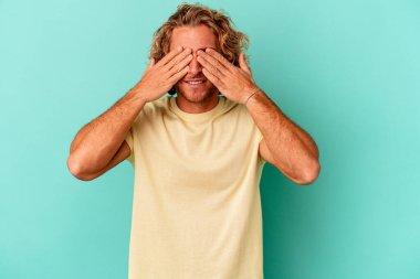 Mavi arka planda izole edilmiş genç beyaz adam elleriyle gözlerini kapatmaktan korkuyor..