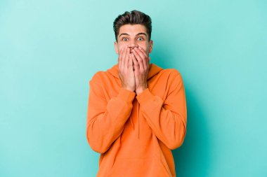 Mavi arka planda izole edilmiş genç beyaz adam şok olmuş, ağzı ellerle kaplanmış, yeni bir şey keşfetmeye hevesli..