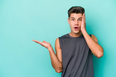 Mavi arka planda izole edilmiş genç beyaz adam, avuç içi fotokopi alanını elinde tutmaktan etkilenmiş..