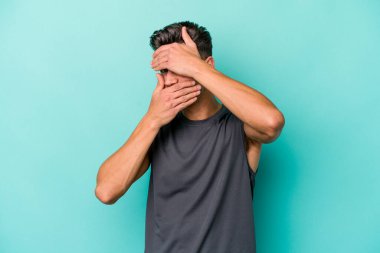 Mavi arka planda izole edilmiş genç beyaz adam kameraya parmaklarıyla göz kırpıyor, yüzünü kapatmaktan utanıyor..