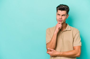 Mavi arka planda izole edilmiş genç beyaz adam şüpheli ve şüpheci bir ifadeyle yana bakıyor..