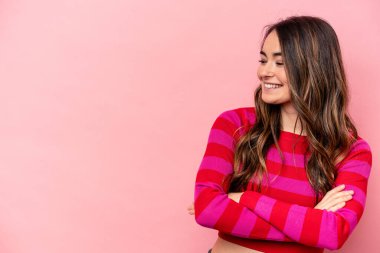 Pembe arka planda izole edilmiş genç beyaz kadın kollarını kavuşturarak gülümsüyor..