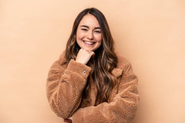 Beyaz tenli genç bir kadın bej arka planda izole edilmiş mutlu ve kendinden emin gülümsüyor, elleriyle çenesine dokunuyor..