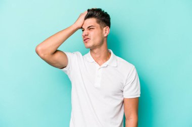 Kafkasyalı genç adam mavi arka planda izole edilmiş bir şekilde bir şeyi unutuyor. Alnına avuç içi ve gözlerini kapayarak vuruyor..