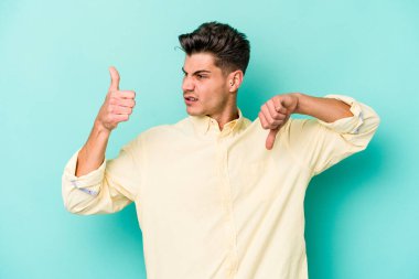 Kafkasyalı genç adam mavi arka planda izole edilmiş, baş parmağını kaldırıyor, zor seçim konsepti.