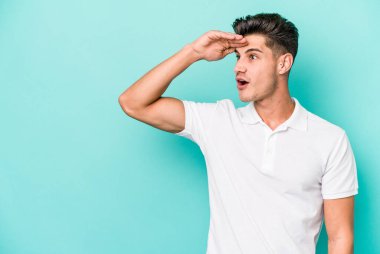 Kafkasyalı genç adam mavi arka planda izole edilmiş, uzaklara bakıyor ve elini alnına koyuyor..