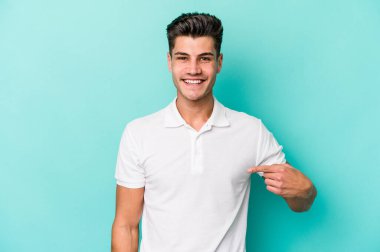 Mavi arka planda izole edilmiş genç beyaz adam bir gömlek fotokopi alanını işaret ediyor, gururlu ve kendinden emin.