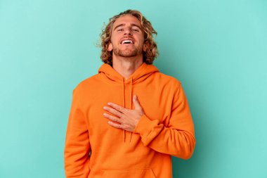 Mavi arka planda izole olmuş genç beyaz adam yüksek sesle gülüyor ve elini göğsüne koyuyor..
