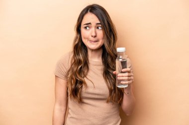 Kafkasyalı genç bir kadın bej arka planda izole edilmiş bir şişe su tutuyor. Kafası karışmış ve kendinden emin değil..