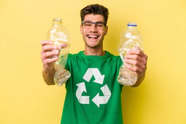 Young caucasian man holding a bottle of plastic to recycle isolated on yellow background