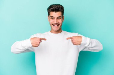 Mavi arka planda izole edilmiş genç beyaz adam parmağını göstererek şaşırdı, geniş bir gülümsemeyle..