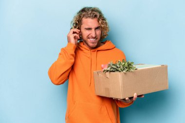 Young caucasian man making a move while picking up a box full of things isolated on blue background covering ears with hands.