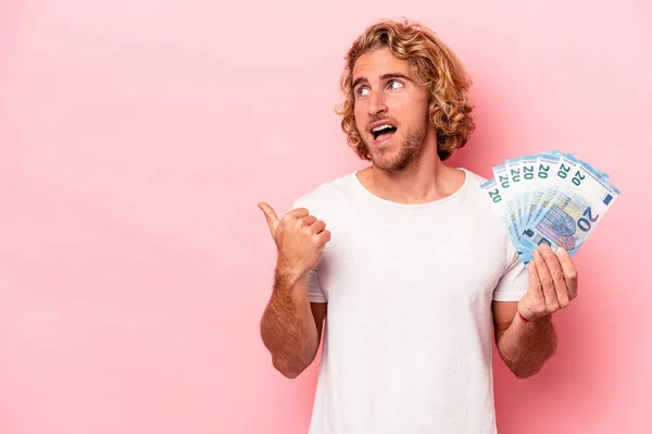 Young caucasian man holding banknotes isolated on pink background points with thumb finger away, laughing and carefree.