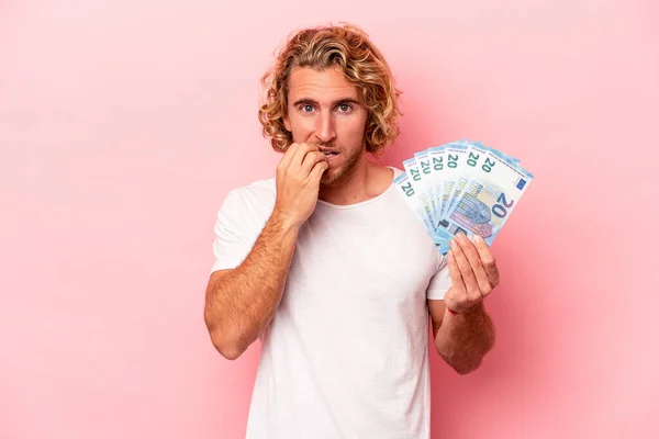 Young caucasian man holding banknotes isolated on pink background biting fingernails, nervous and very anxious.