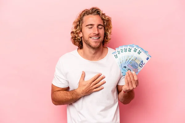 Young caucasian man holding banknotes isolated on pink background laughs out loudly keeping hand on chest.