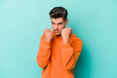 Kafkasyalı genç bir adam mavi arka planda izole edilmiş. Yumruk atıyor, sinirleniyor, kavga ediyor, kavga ediyor..