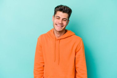 Mavi arka planda izole edilmiş genç beyaz adam gülüyor ve gözlerini kapıyor, rahatlamış ve mutlu hissediyor..